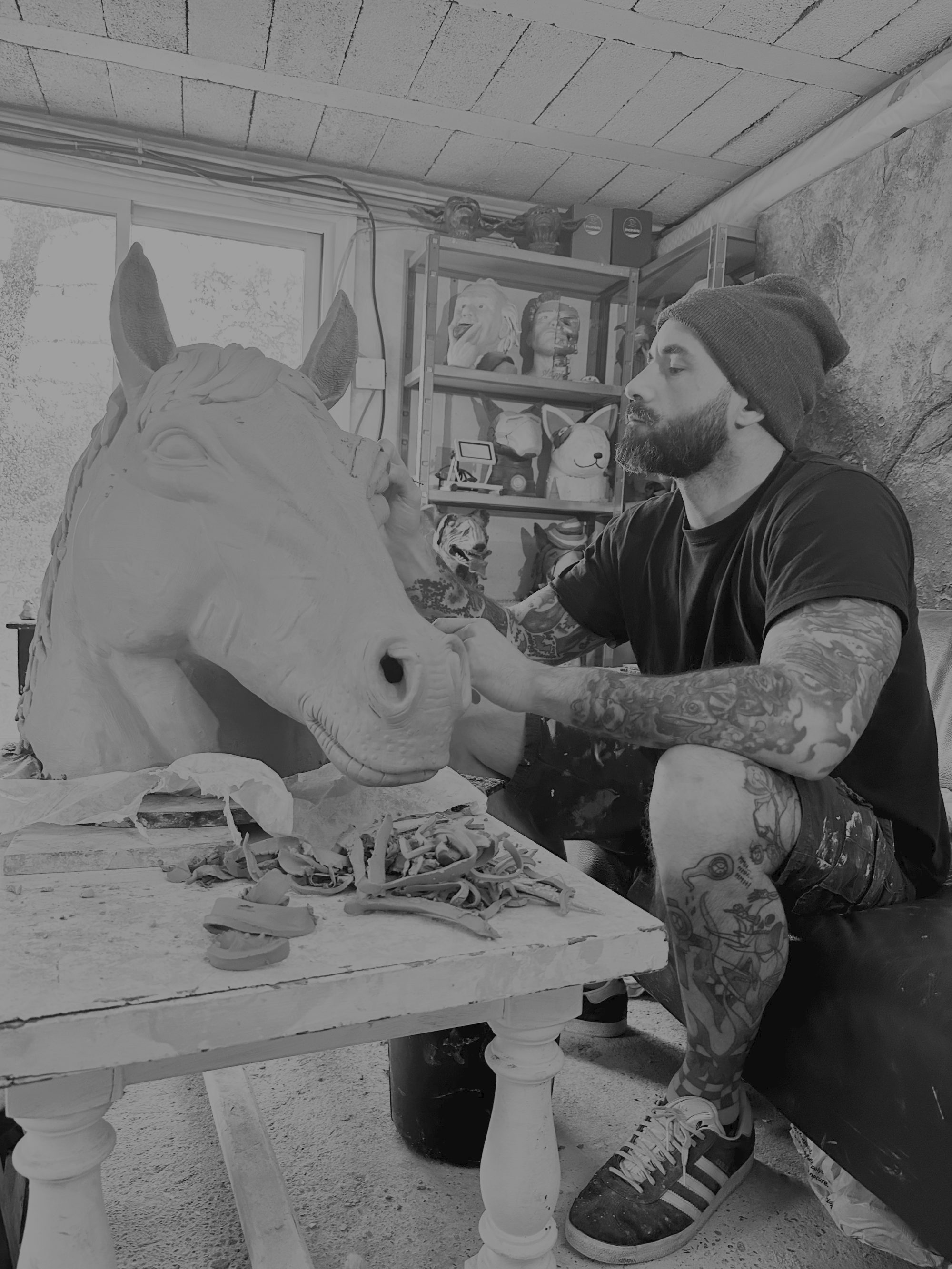 Dyson, de profil, façonne à la main une grande tête de cheval en argile dans son atelier ; derrière lui, des étagères de têtes sculptées.